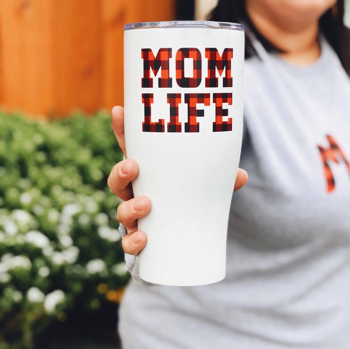 Someone holds the Mom Life Plaid • Tumbler, featuring bold red and black buffalo plaid letters. The stainless steel travel mug stands out against a blurry green and wood background, while the person wears a light gray shirt.