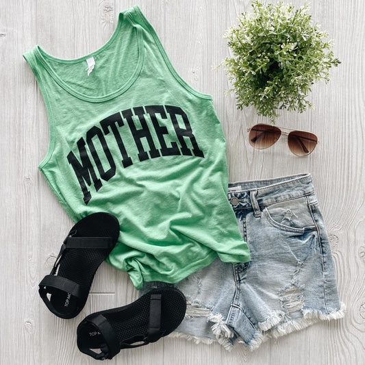 A flat lay displays the MOTHER • Unisex Tank Top in green, styled with distressed denim shorts, black sandals, brown sunglasses, and a small potted plant on a light wood surface. Features college block letters for effortless style.