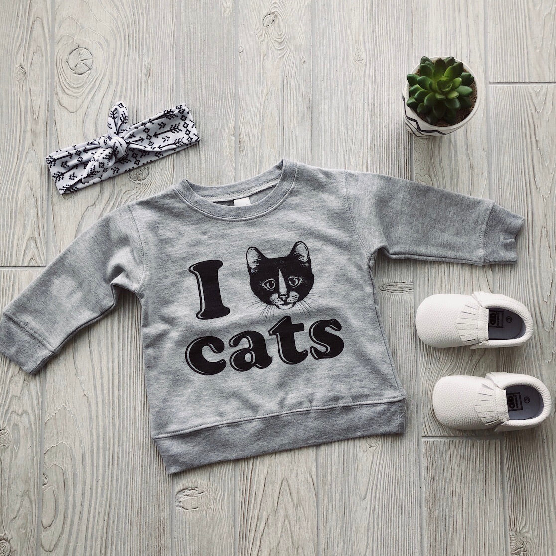 The I Love Cats • Pullover, a gray childrens sweatshirt with an adorable cat face print, is shown with a patterned headband, white baby shoes, and a small potted succulent on a wooden surface.