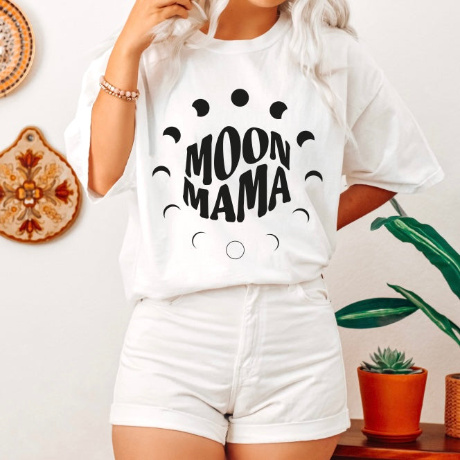 A person with long, light hair wears the MOON MAMA • Comfort Colors Tee featuring moon phases, paired with white shorts. The background has wall décor, a green plant, and a small succulent.