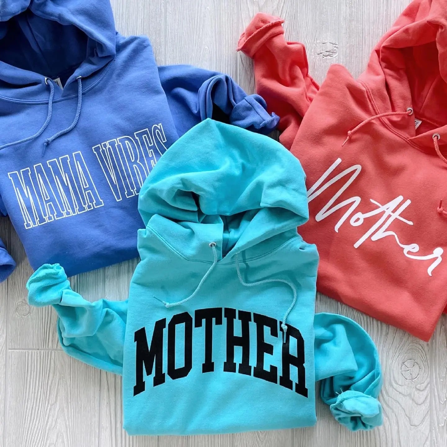 The MOTHER • Aqua Hoodie, featuring bold black MOTHER lettering, is laid out on a light wooden floor alongside blue and coral sweatshirts with white lettering.