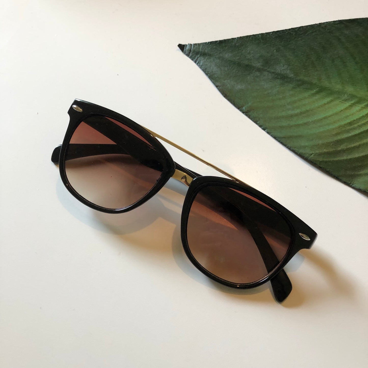 A pair of Wayfarer Bar Sunglasses for adults, featuring black frames and UV400 dark lenses, rests on a white surface beside a green leaf-shaped object.