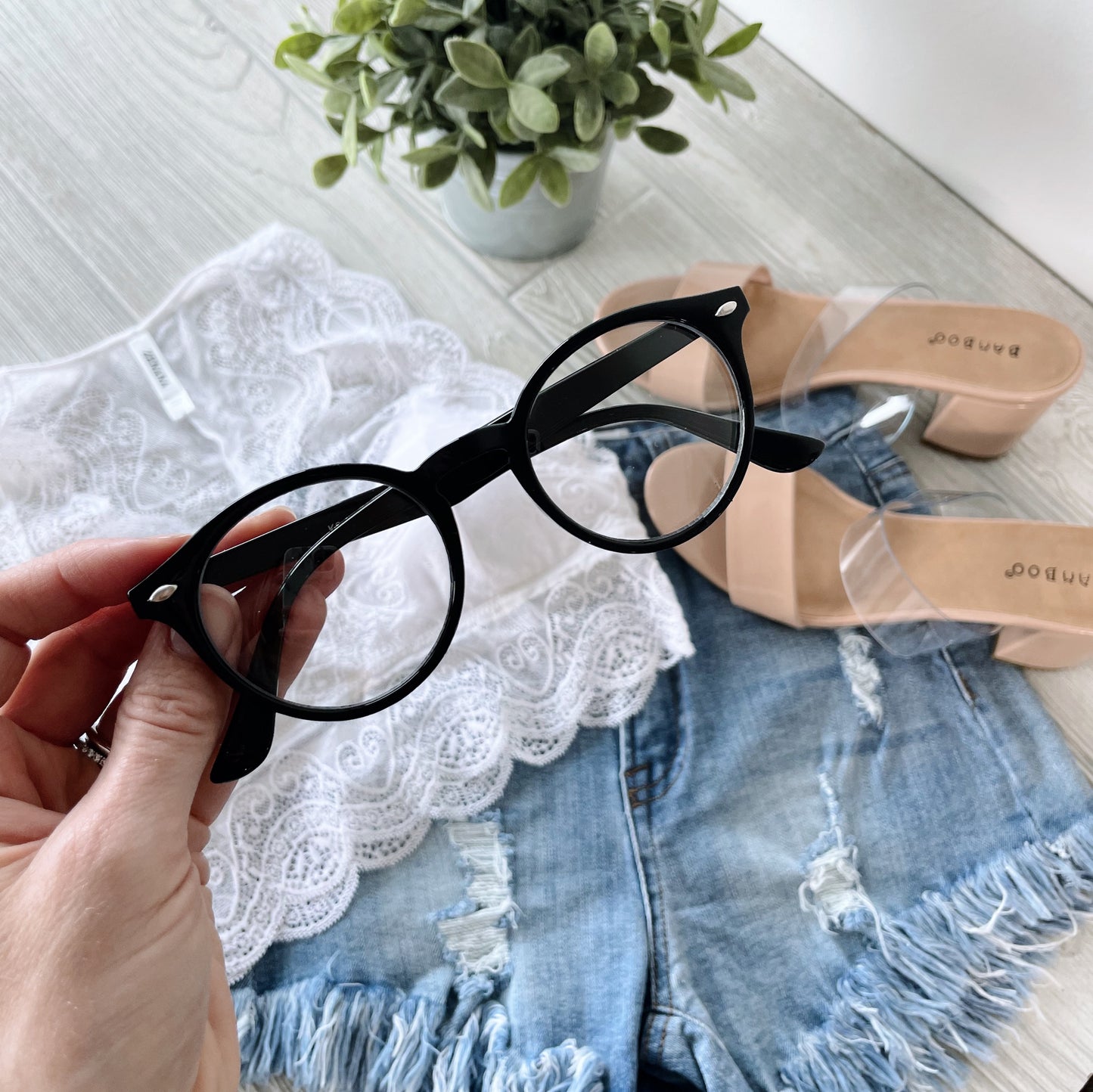 A hand holds Harper Clear Lens • Adult glasses above a flat lay featuring denim shorts, a white lace top, nude heeled sandals, and a small potted plant on a light wooden surface.