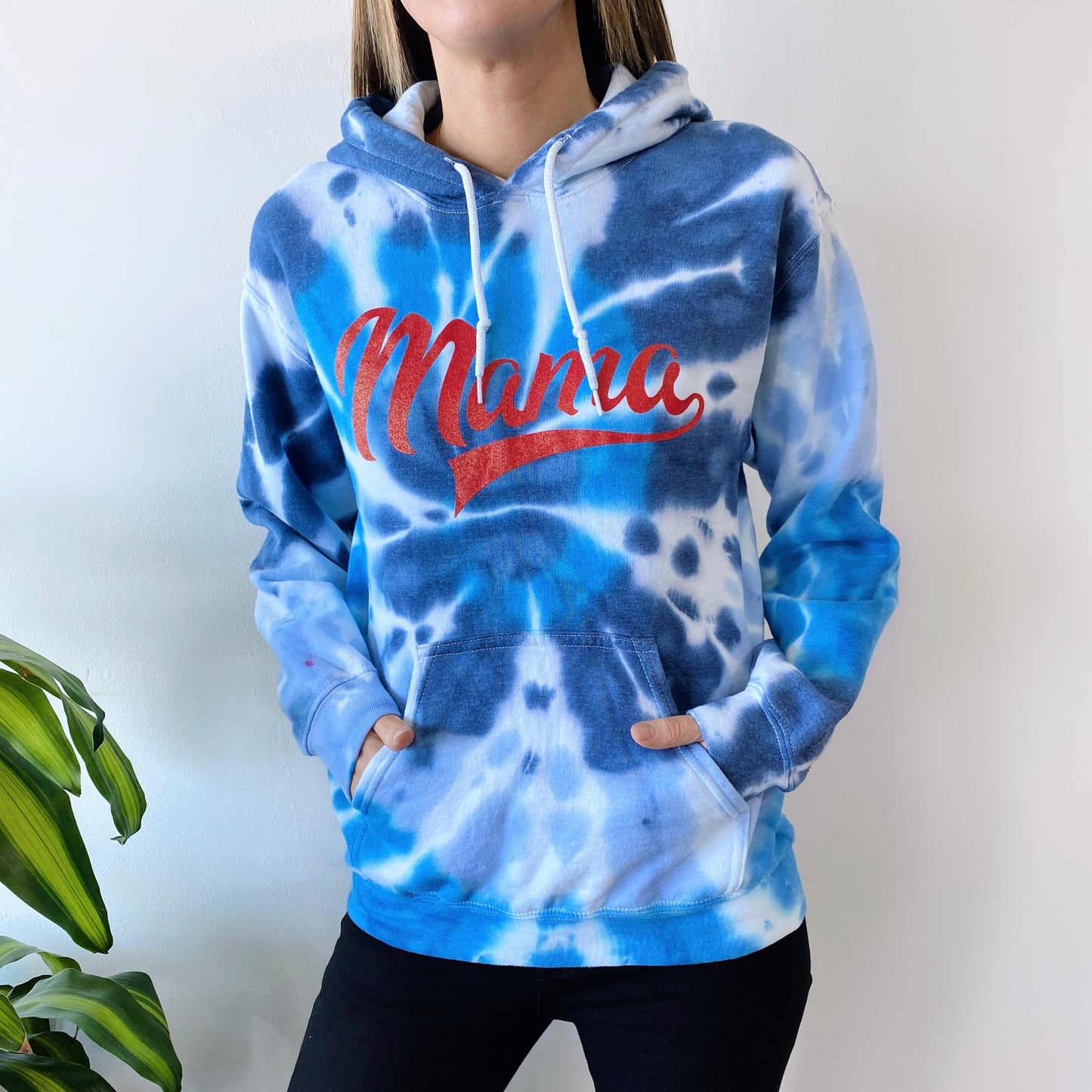 A person wears the Americana • Tie-Dye Hoodie—a blue and white fleece pullover with Mama in bold red script—standing against a plain white background. A green plant on the left adds a fresh accent to this sporty, stylish look.