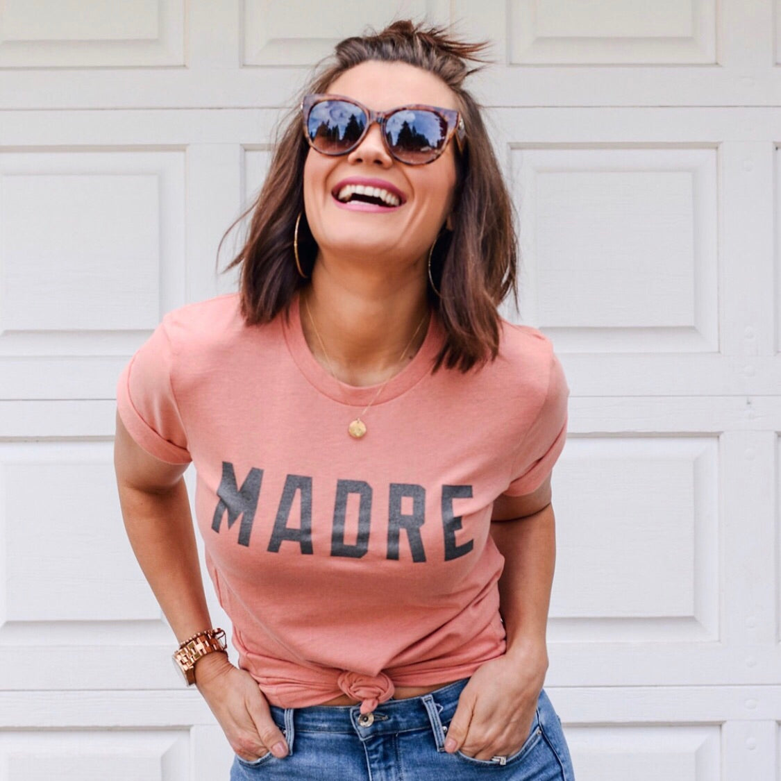 A smiling woman with shoulder-length brown hair and gold hoop earrings wears gunmetal sunglasses, jeans, and the Madre Tee in Spring Coral (Bella Canvas, MADRE print) while standing in front of a white garage door.