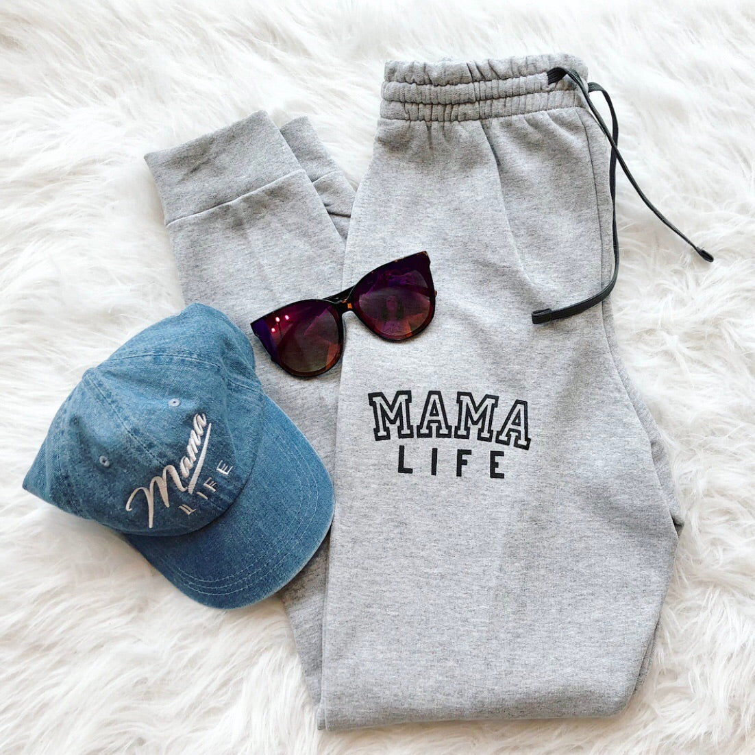 The Mama Life Joggers in gray, styled with dark sunglasses and a blue Mama Life embroidered cap, are displayed on a white, fluffy surface for the ultimate cozy loungewear vibe.