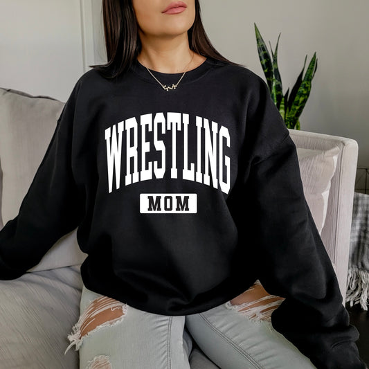 A woman with long brown hair sits on a couch wearing the Wrestling Mom • Black Pullover, featuring bold white lettering. She pairs it with ripped light gray jeans, and a green plant is visible in the background.