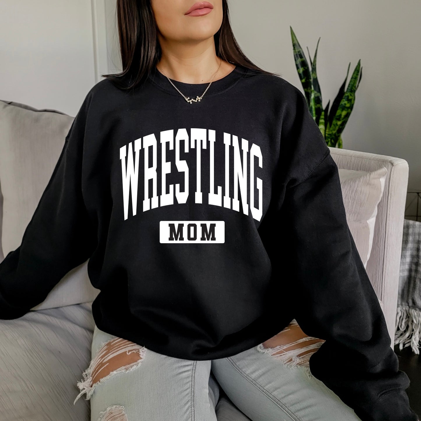 A woman with long brown hair sits on a couch wearing the Wrestling Mom • Black Pullover, featuring bold white lettering. She pairs it with ripped light gray jeans, and a green plant is visible in the background.