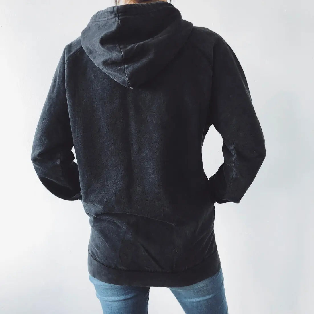 A person stands with their back to the camera, wearing the MAMA VIBES • Rocker Hoodie in black vintage wash and blue jeans against a plain white background.