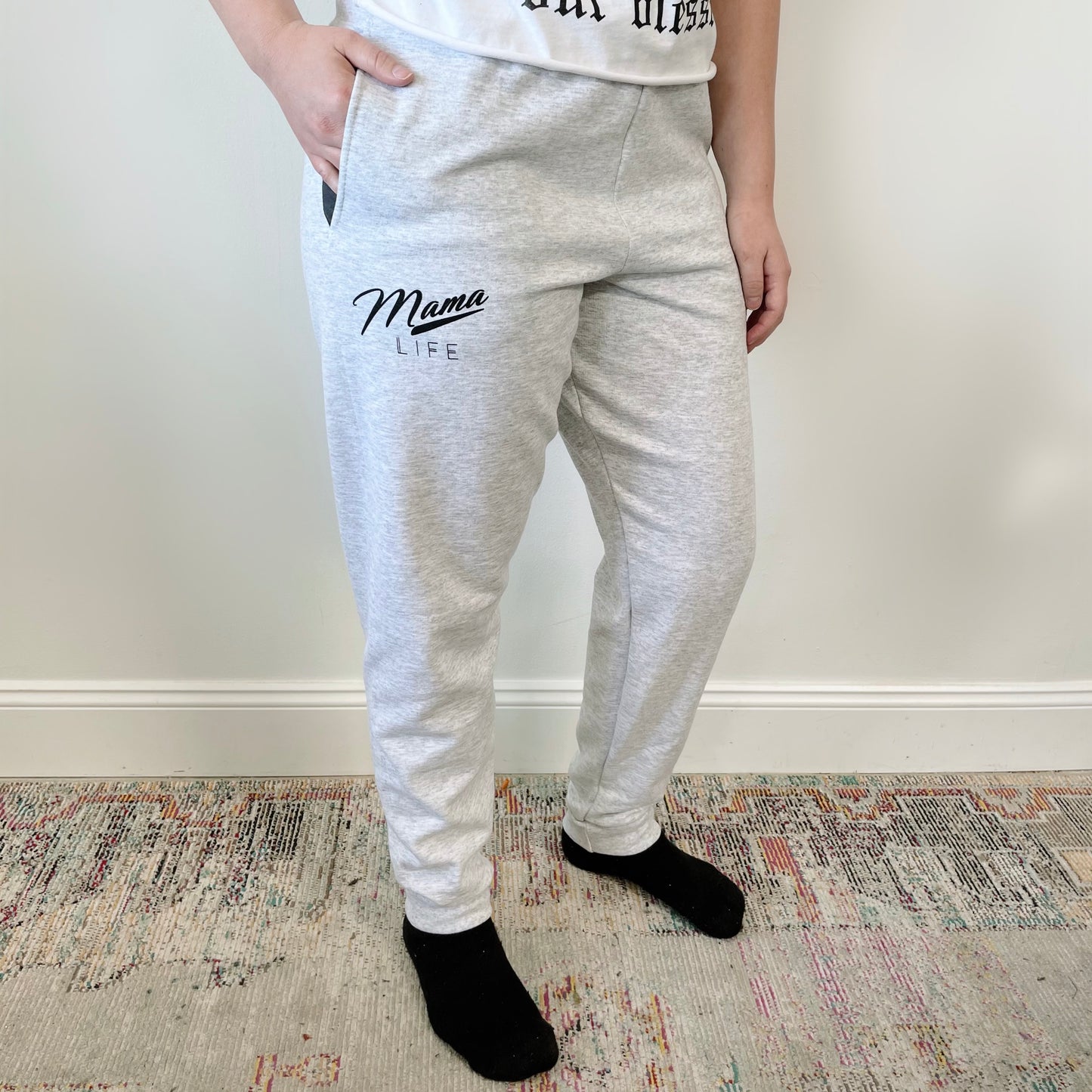 A person wearing Mama Retro • Ash Joggers, featuring a retro Mama Life print on light gray fabric, paired with black socks and a white shirt, stands on a patterned rug against a plain white wall for an effortless jogger style.
