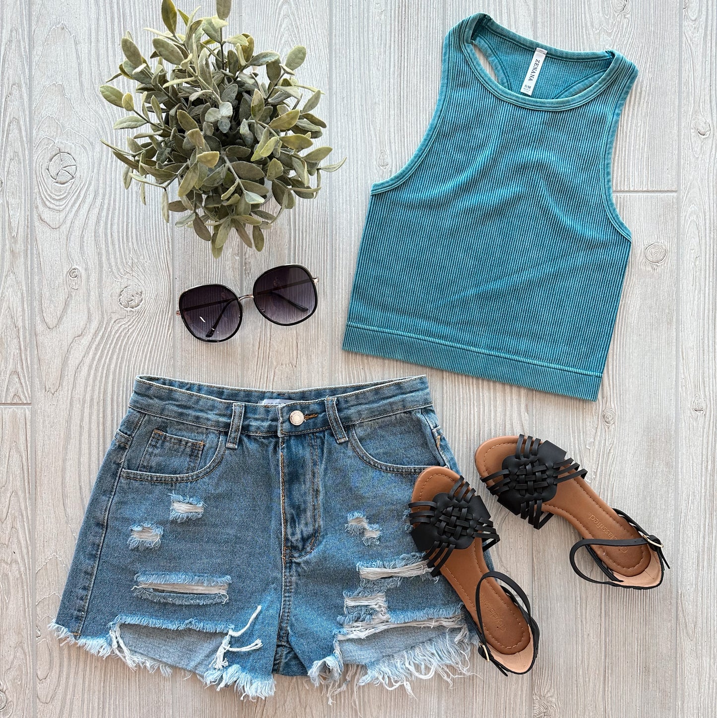 Flat lay of a casual summer outfit featuring the Sophia High-Neck Crop in vintage teal, distressed denim shorts, black slides with decorative detail, dark sunglasses, and a small potted plant on a light wooden surface.