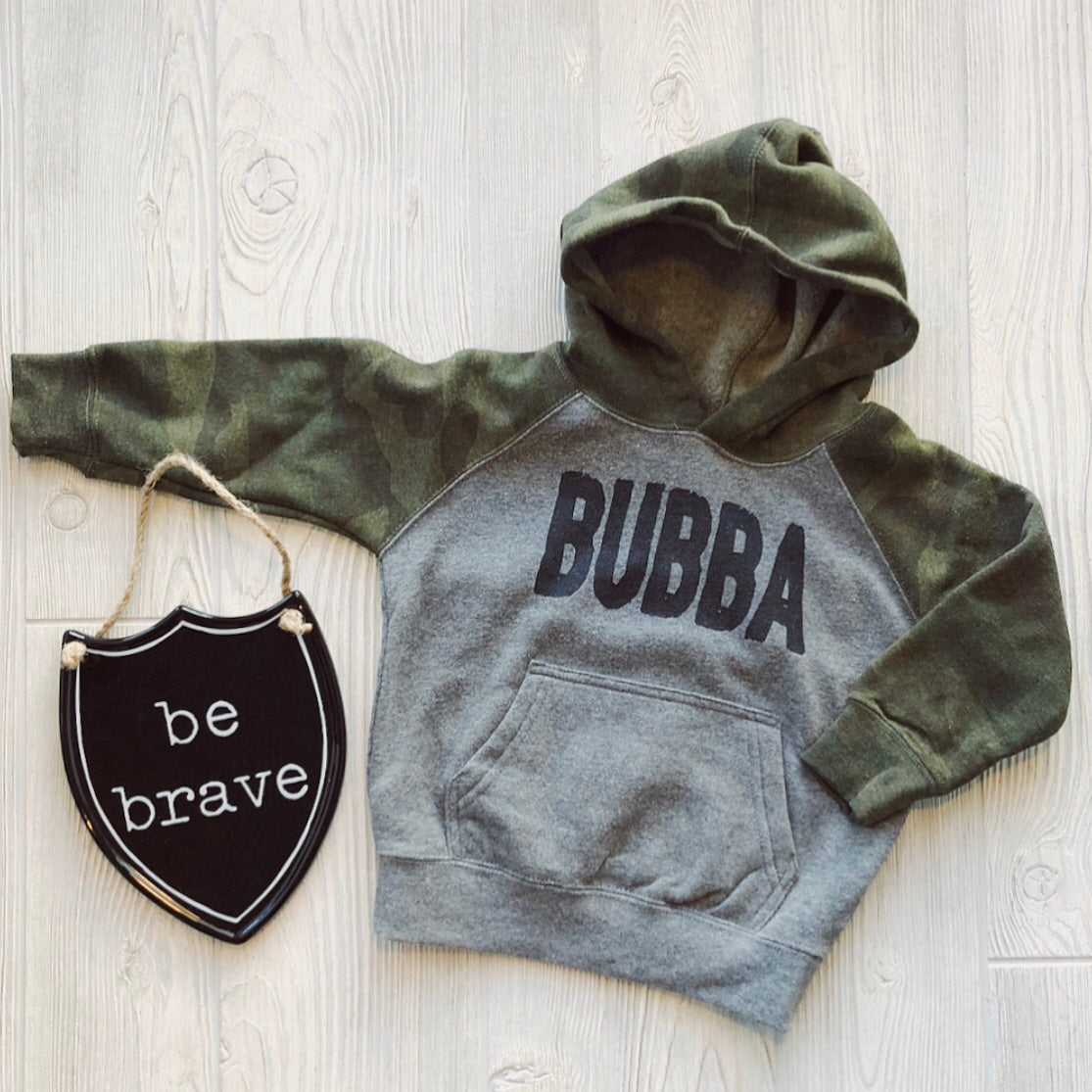 The BUBBA • Camo Sleeve Hoodie—gray with green camo sleeves and BUBBA on the front—is paired with a black shield sign reading be brave in white, both shown on a light wooden surface. Ideal for your childs spring wardrobe.
