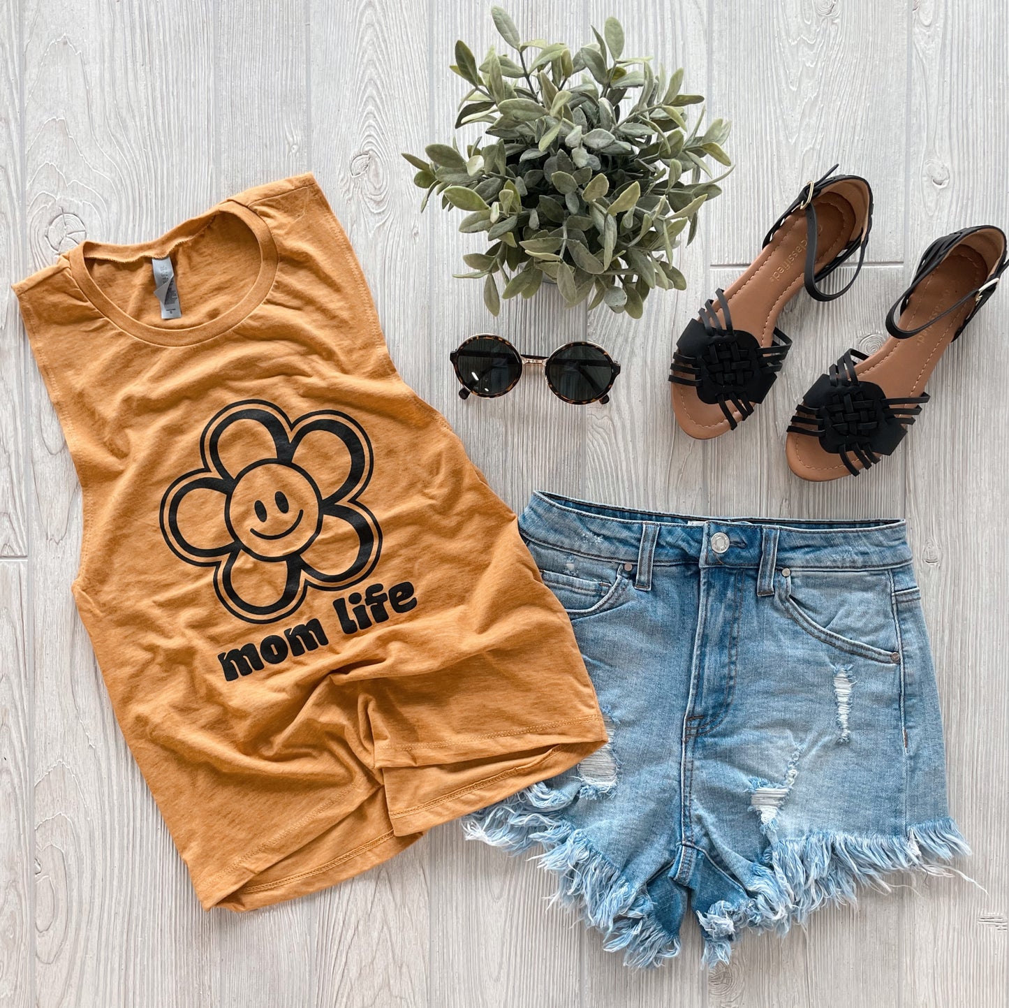 Flat lay of the Happy Flower • Tank, featuring a groovy flower print and mom life text in mustard yellow, styled with distressed denim shorts, black sunglasses, black sandals, and a small potted plant on a light wood floor.