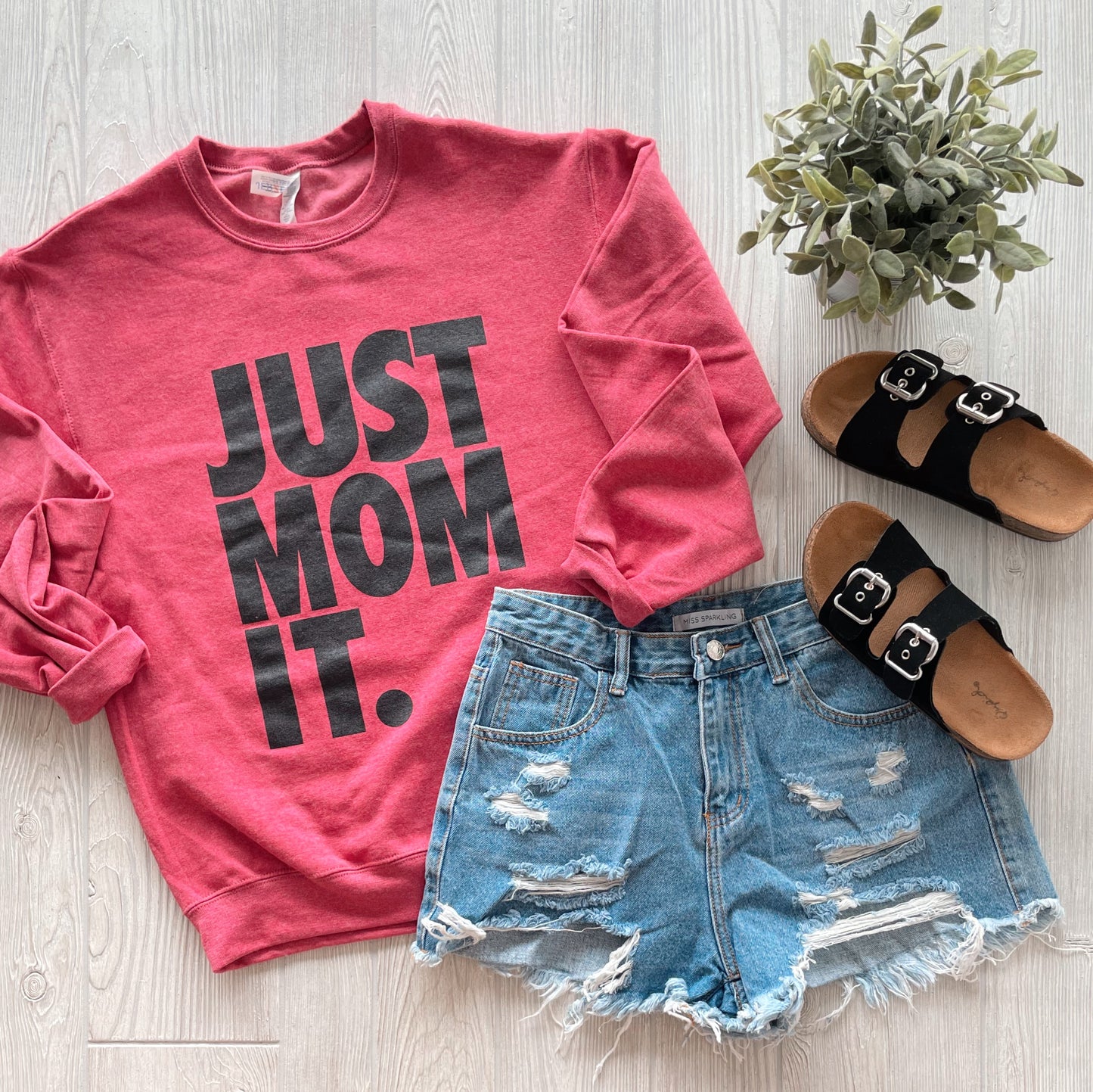 A flat lay of the JUST MOM IT • Heather Red Pullover with bold black lettering, paired with distressed denim shorts, black buckle sandals, and a small potted plant on a light wood surface.