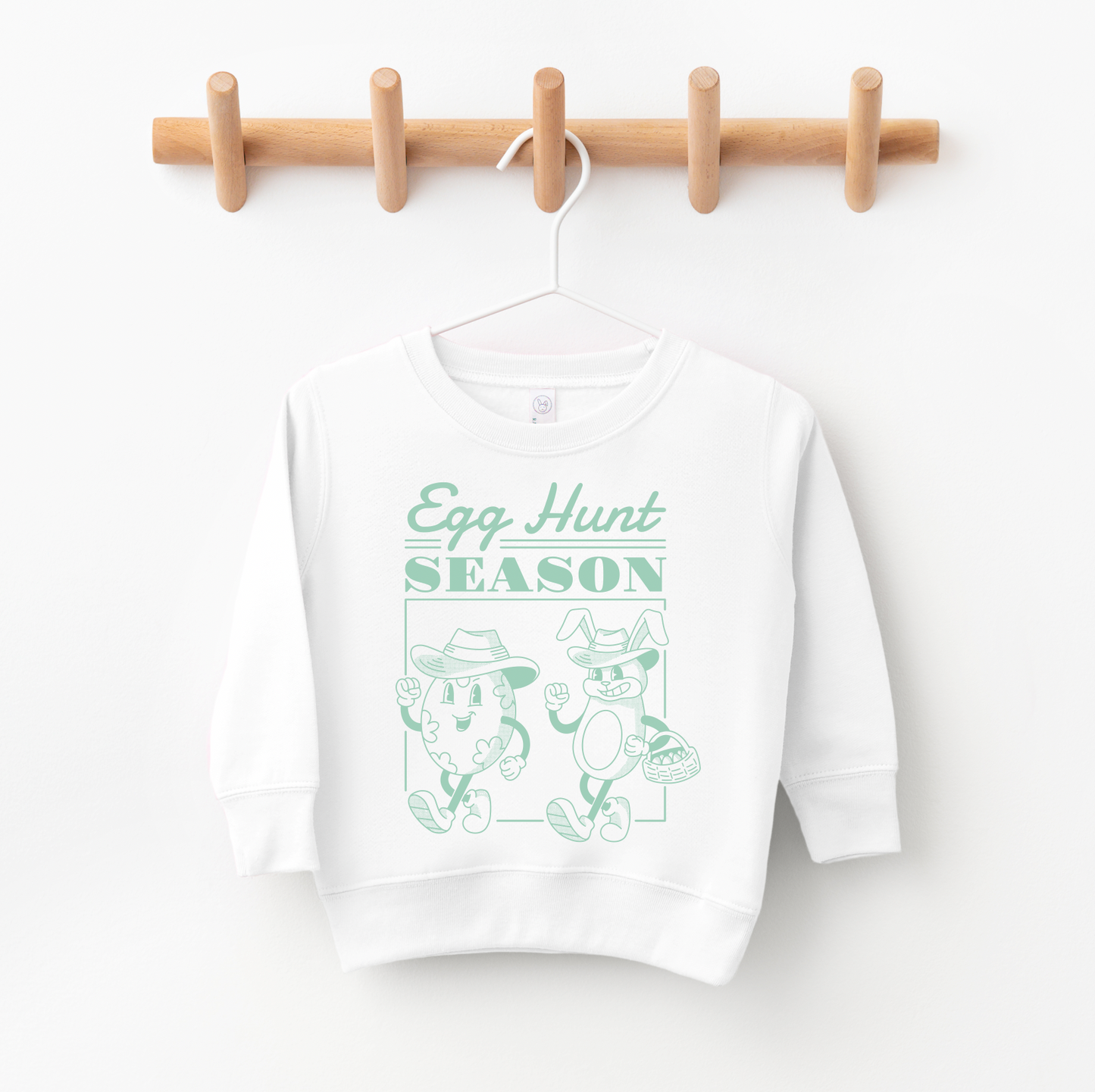 The Egg Hunt Pullover • Youth or Adult in white features a mint green line drawing of two eggs in hats with baskets and Egg Hunt SEASON text. Shop small for this playful, unisex spring essential!.