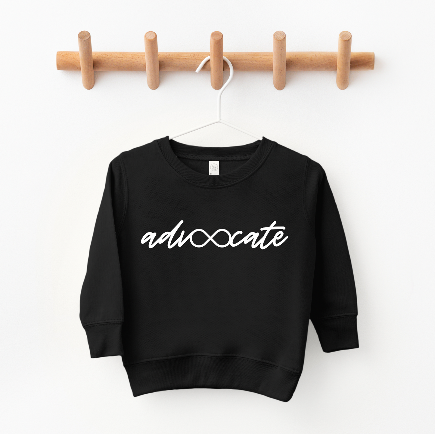 The Advocate • Kids Pullover is a black, oversized-fit sweatshirt with advocate in white script and an infinity symbol for the vo, displayed on a wooden peg rack against a white wall—perfect for any kids clothing collection.