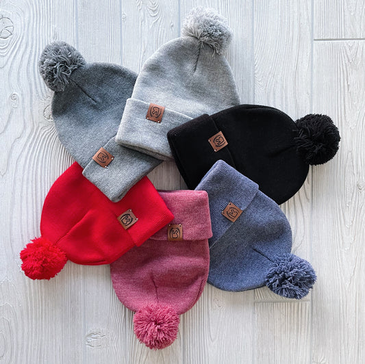 Six Pom Collection Beanies in gray, black, red, pink, and blue—each hand-sewn with a pom-pom and leather logo patch—are displayed fan-style on a light wood surface.
