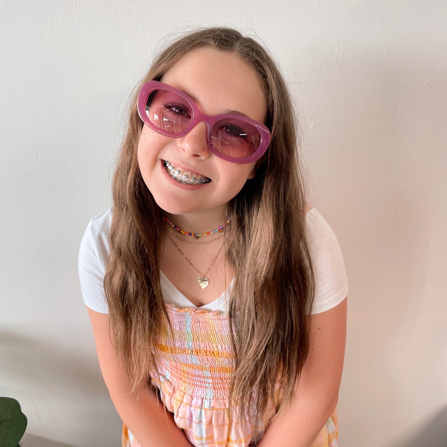 A smiling girl with long brown hair wears Poppy Sunglasses (adult size, UV protected), braces, layered necklaces, a white t-shirt, and a colorful dress while standing in front of a plain white wall.