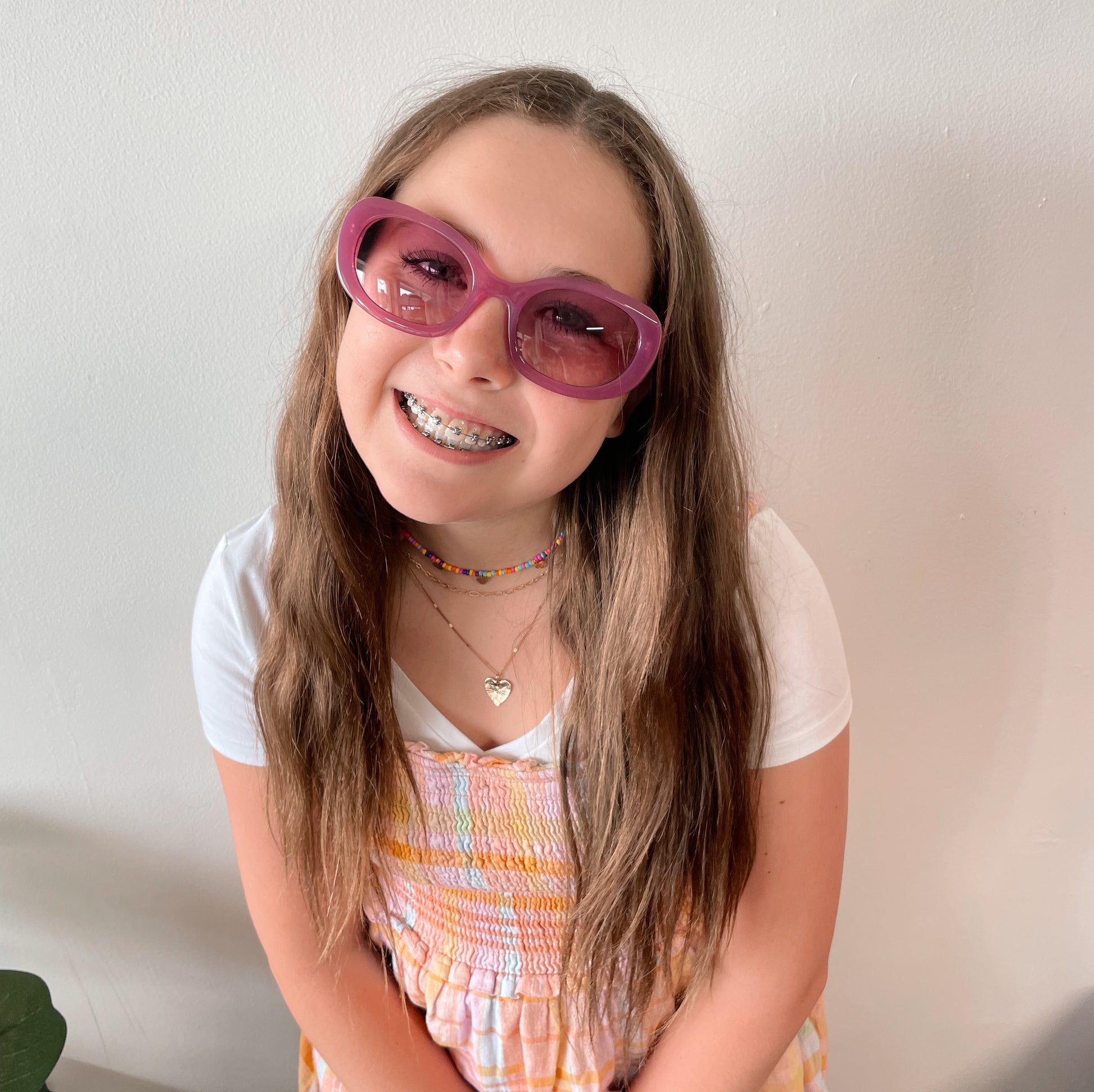 A smiling girl with long brown hair wears Poppy Sunglasses (adult size, UV protected), braces, layered necklaces, a white t-shirt, and a colorful dress while standing in front of a plain white wall.