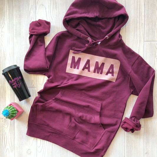 The MAMA Box Shimmer • Maroon Hoodie, featuring bold MAMA text in custom mixed ink, is displayed on a light wooden floor next to a black travel mug with pink writing and a small green cactus in a pink pot.