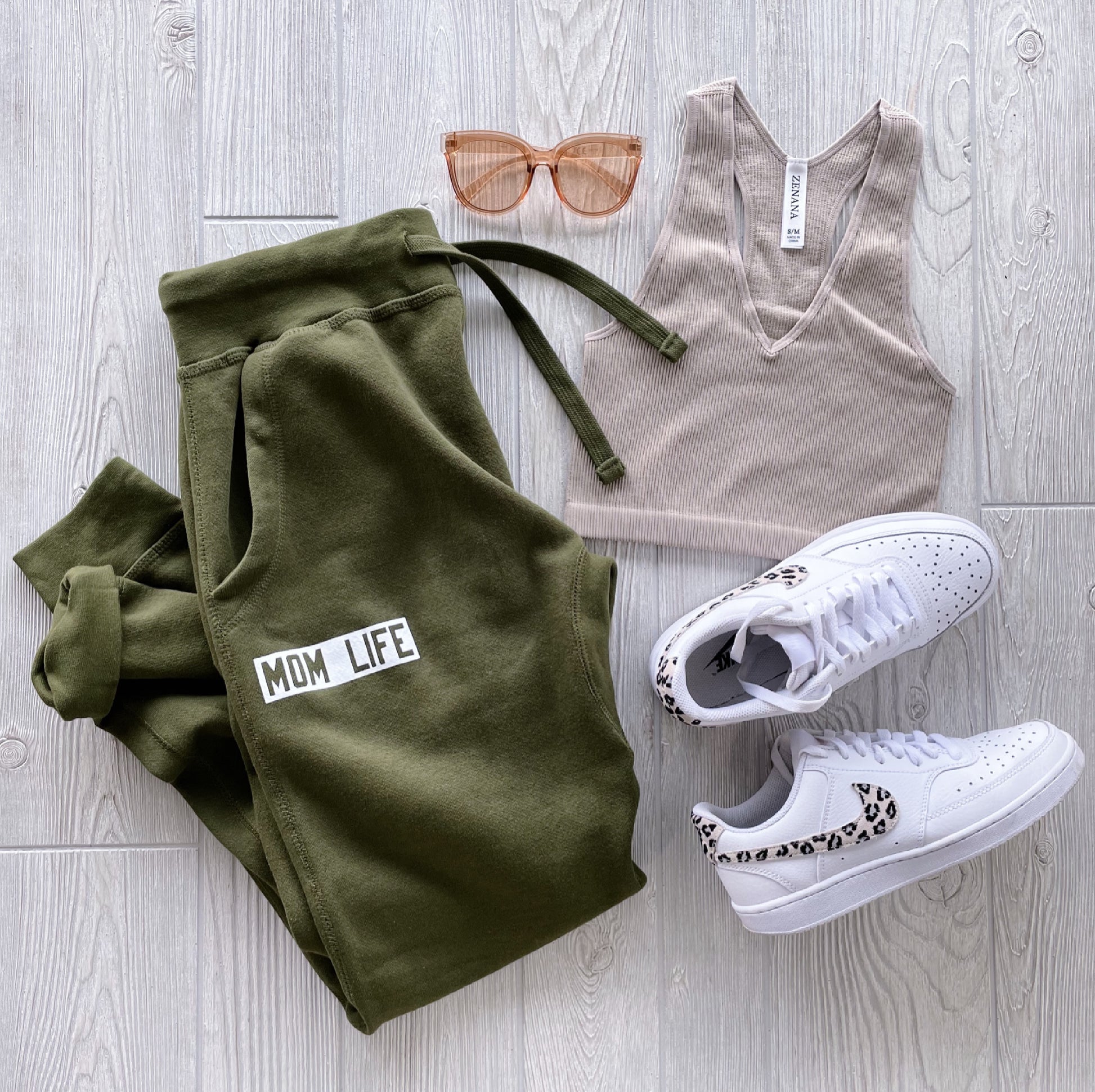 Flat lay of casual loungewear: Mom Life • Olive Joggers, taupe ribbed crop top, white Nike sneakers with leopard print accents, and brown-tinted sunglasses on a light wood floor.