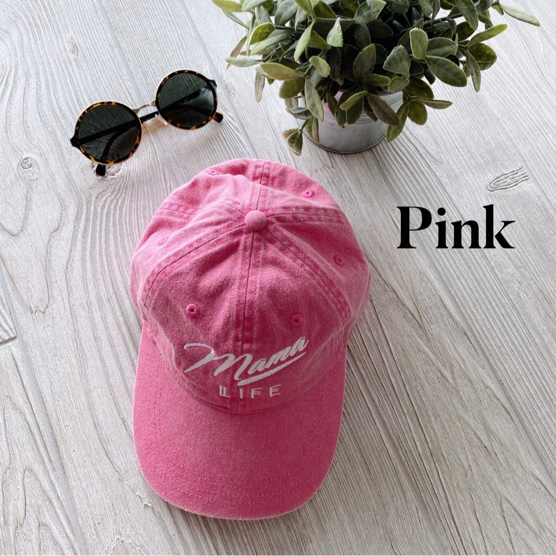 A Mama Life Hat • NEW Colors! in pink, embroidered with care, sits on a light wooden surface next to round sunglasses and a leafy potted plant. The word Pink appears in bold black text on the right side of the image.