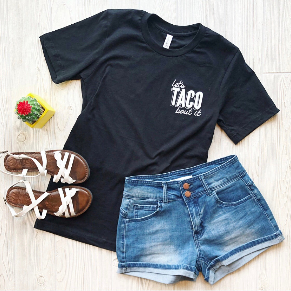 Flat lay of the Taco Tee • Adult! with lets TACO bout it printed in white, styled with denim shorts, white strappy sandals, and a potted cactus—perfect mom tee vibes on a light wooden surface.
