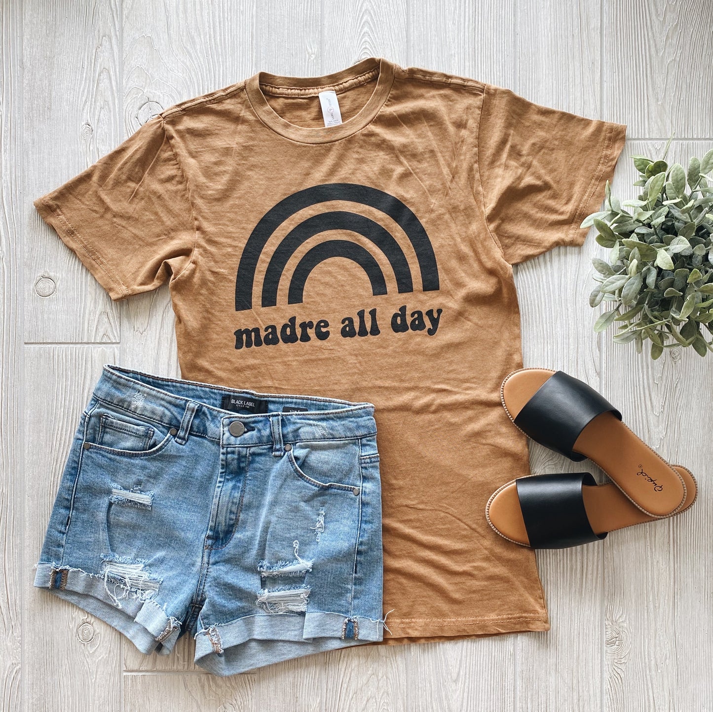 The Madre All Day • Rocker Tee, a tan unisex shirt with a black rainbow print and madre all day text, is styled with distressed denim shorts, black slide sandals, and a small green plant on a light wooden floor.