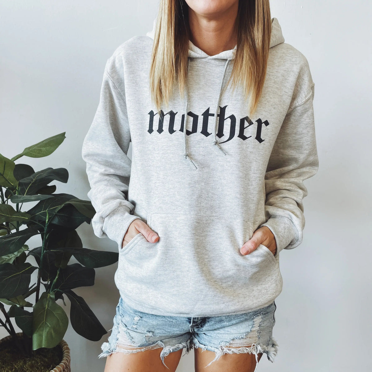 A woman stands by a potted plant with her hands in the pockets of an oatmeal Mother Skull unisex hoodie, made from pill-resistant fleece for lasting comfort. She pairs it with denim shorts. Mother is printed in black on the front.