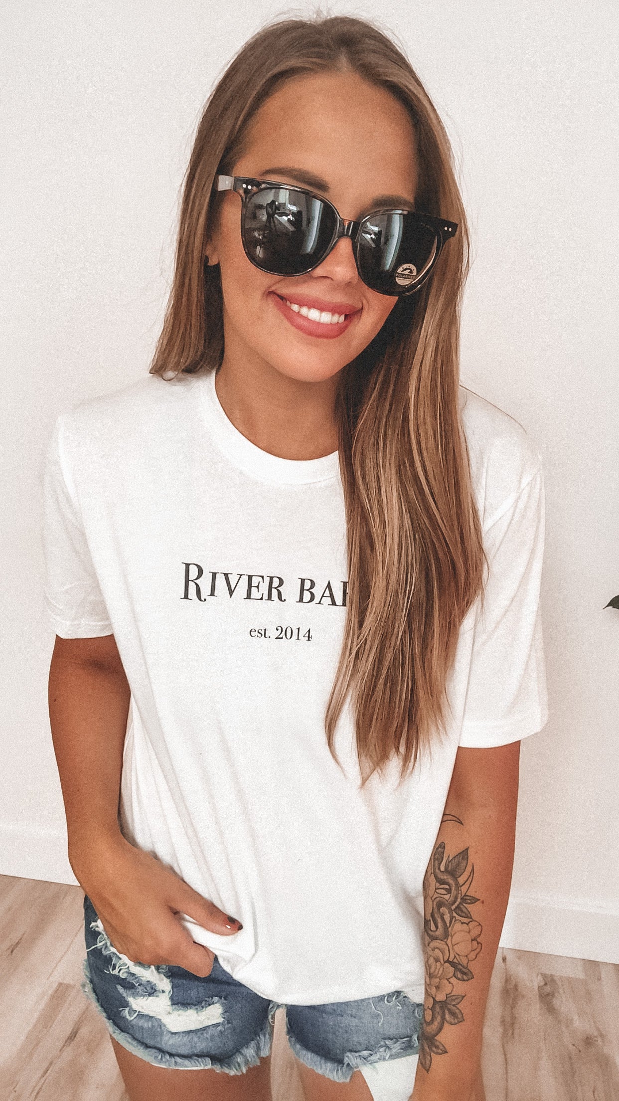 A smiling woman with long hair wears Wayfarer Bar Sunglasses (UV400 Protection, adult size, more colors) and a white River Babe est. 2014 t-shirt. She has a floral tattoo on her left forearm and stands against a light background.
