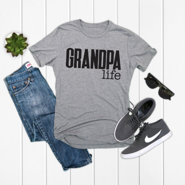 A flat lay of the Grandpa Life • Blue or Gray unisex t-shirt, made from combed and ring-spun cotton, styled with blue jeans, gray Nike sneakers, black sunglasses, and a green succulent on a white wooden background.