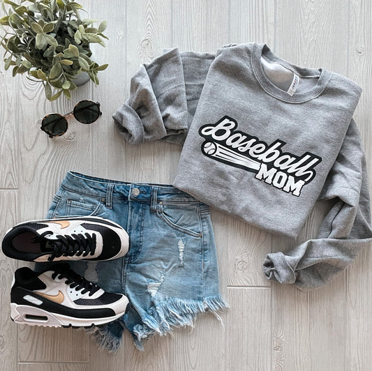 A flat lay of the Baseball Mom • Gray Pullover, distressed denim shorts, black and white sneakers, black sunglasses, and a small green potted plant arranged on a light wooden surface.