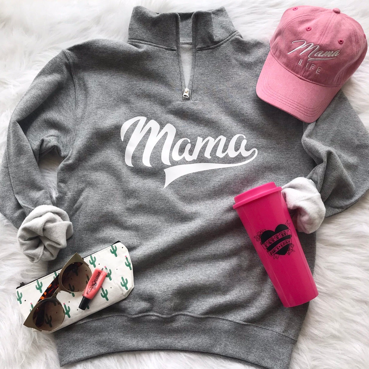 Flat lay of a gray Sporty Mama • Quarter Zip Pullover, pink Mama Life baseball cap, pink tumbler, sunglasses with cactus case, and pink lipstick on a white fluffy surface. This pullover features pill-resistant fleece and a unisex fit.