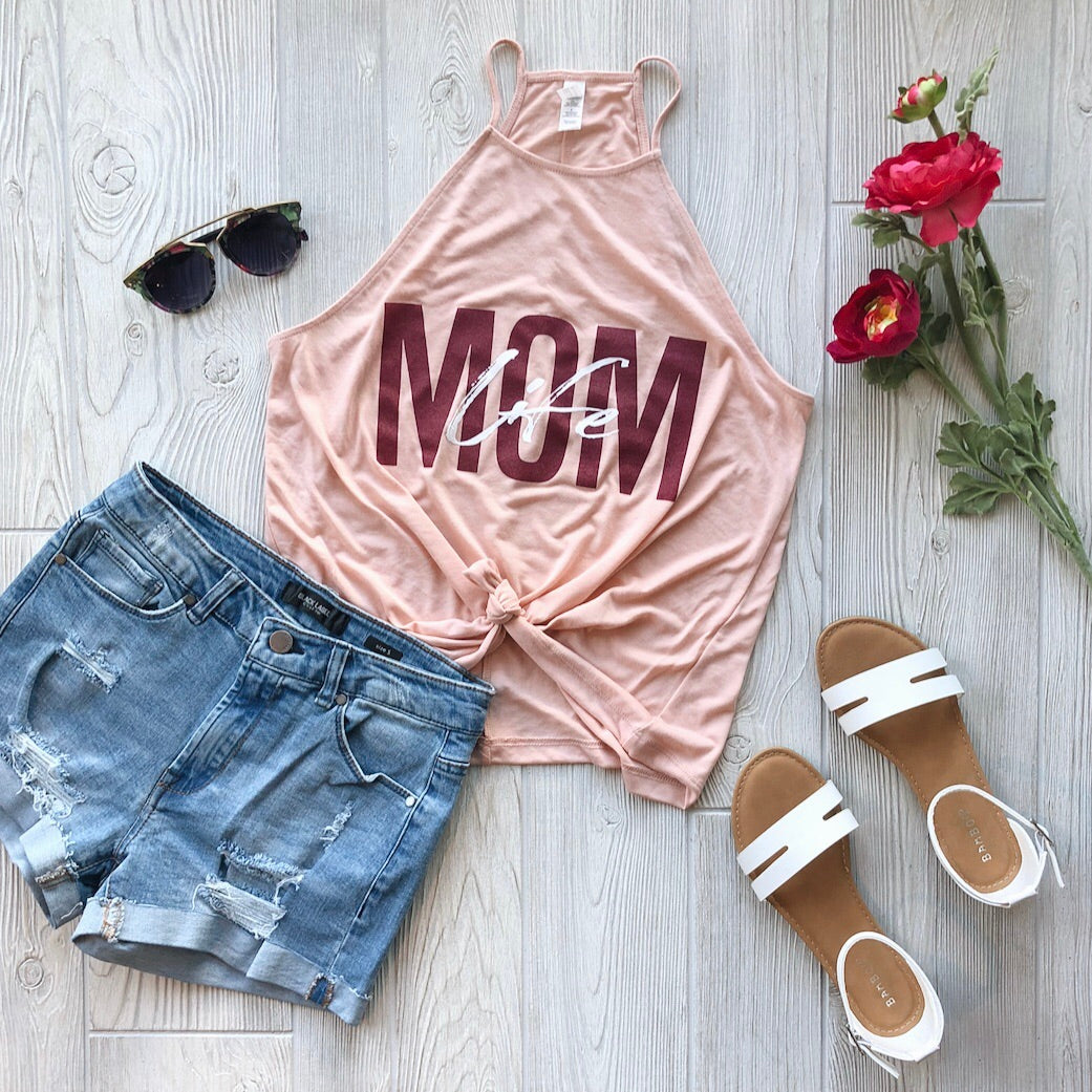 Flat lay featuring the Mom Life Overlay Peach • High Neck Tank, paired with distressed denim shorts, white sandals, black sunglasses, and red flowers on a light wooden floor—perfect for stylishly celebrating mom life.