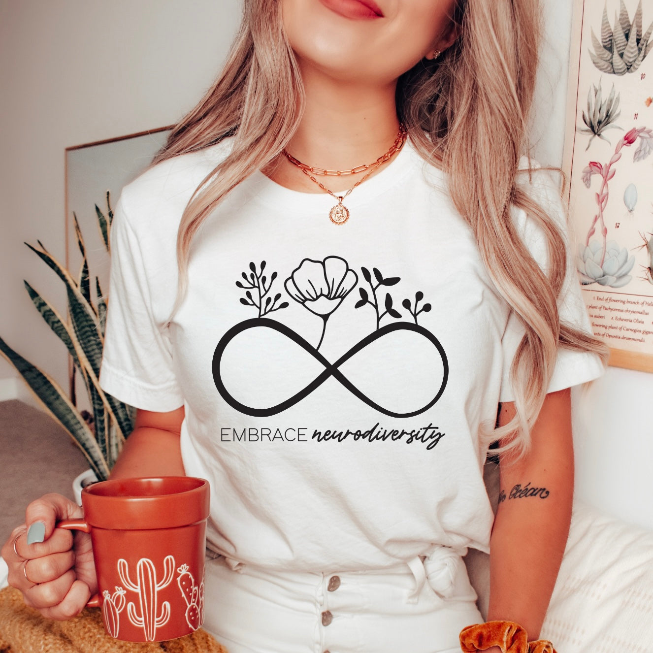 Wearing the Embrace Neurodiversity • White Tee with an infinity symbol and flowers, a person holds a rust cactus mug in a cozy plant-filled room. The shirt has a modern retail fit and promotes neurodiversity.