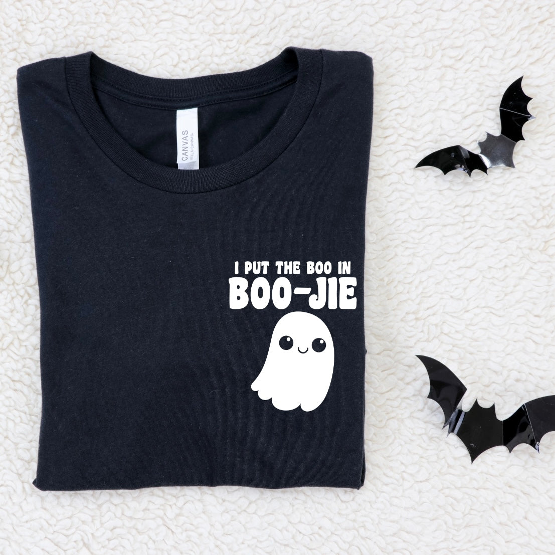 The Boo in BOO-JIE • Black Tee is a unisex black cotton Bella Canvas shirt featuring a cute ghost and “I put the BOOJIE in boo-jie” on the front, displayed folded on a white textured surface beside two black paper bats.