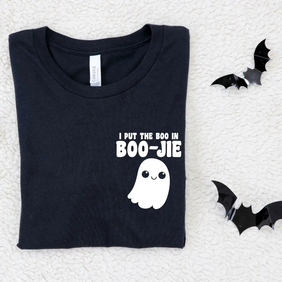 The Boo in BOO-JIE • Black Tee is a unisex black cotton Bella Canvas shirt featuring a cute ghost and “I put the BOOJIE in boo-jie” on the front, displayed folded on a white textured surface beside two black paper bats.