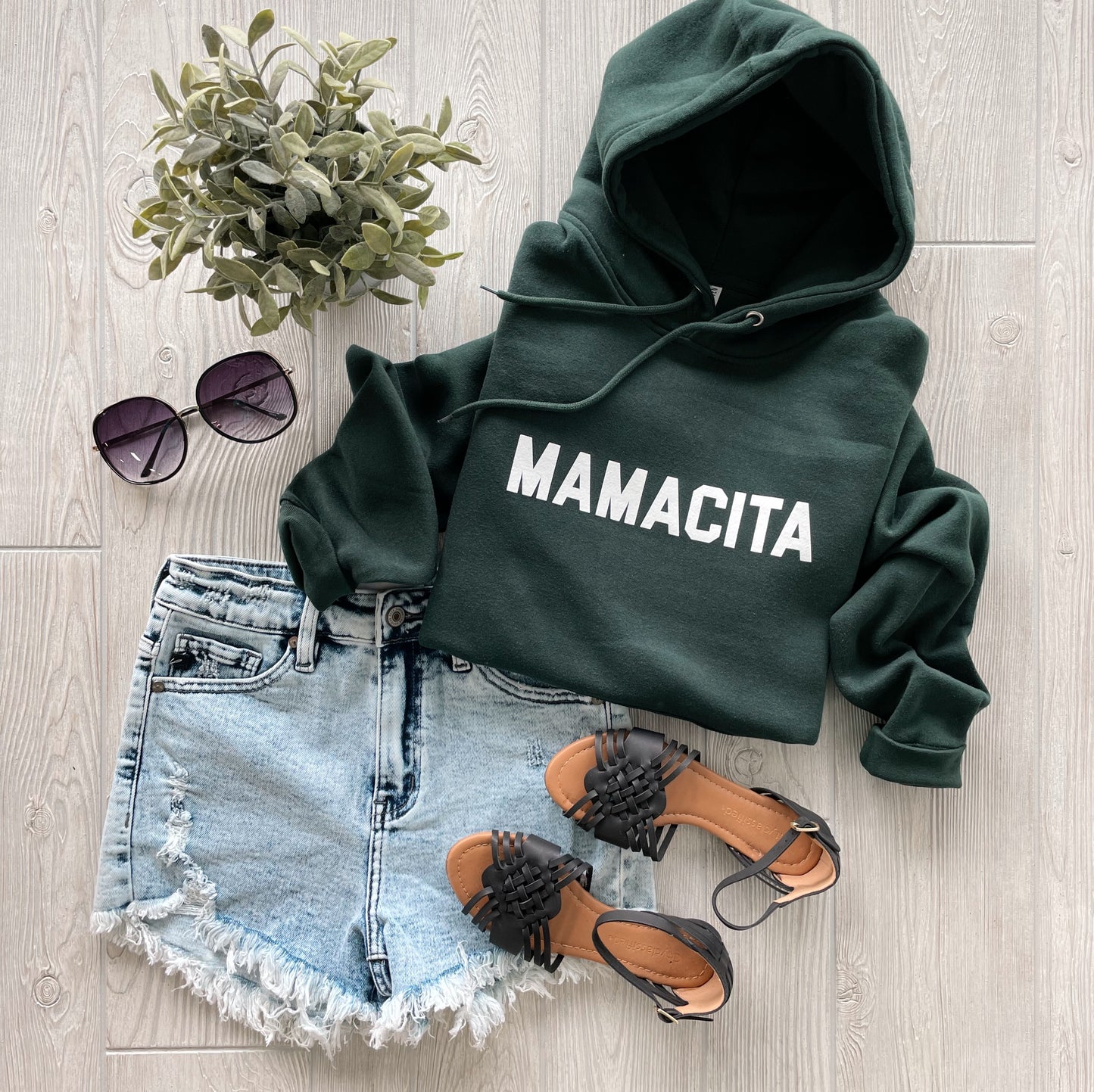 A flat lay featuring the MAMACITA • Sports Green Hoodie with white print, styled with distressed denim shorts, black woven sandals, round sunglasses, and a small green potted plant on a light wood floor.