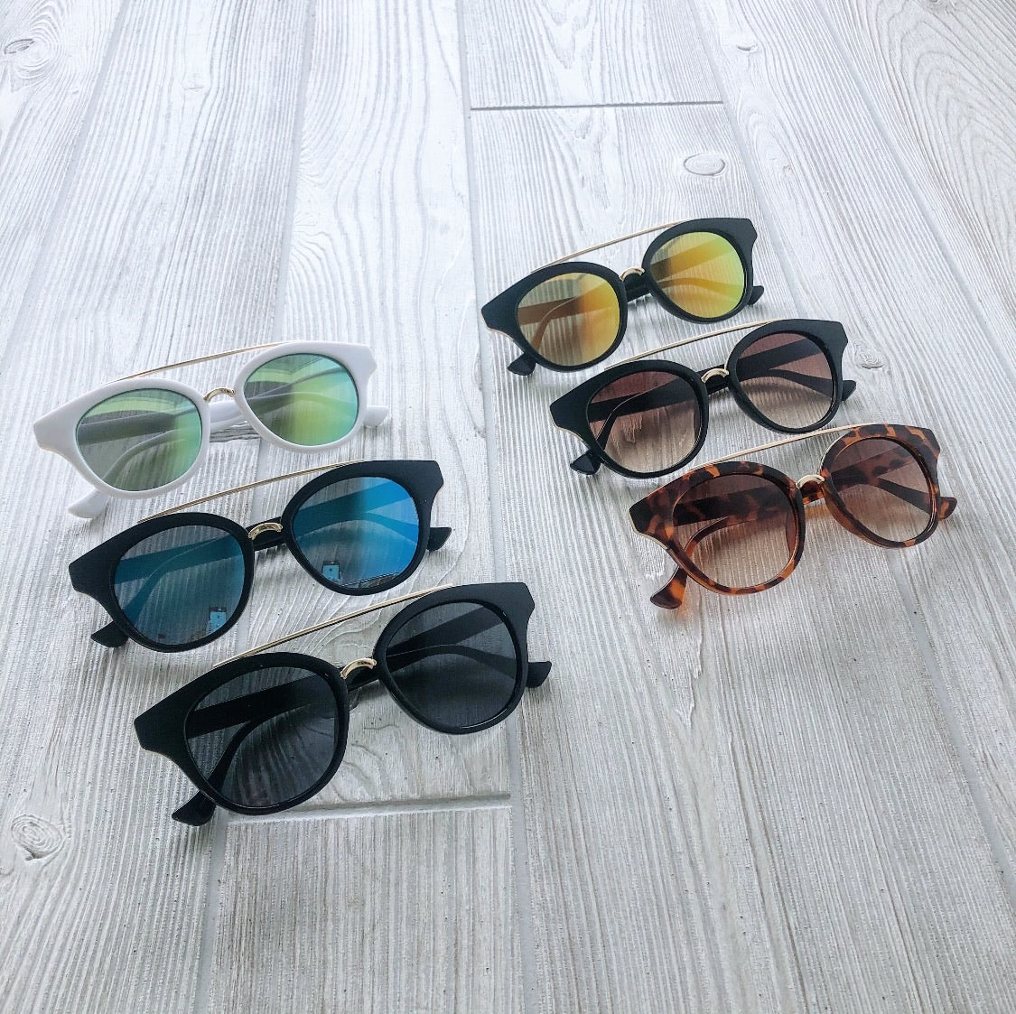 Six pairs of Angie • Sunglasses Glasses • More Colors! are arranged on a light wood floor, showcasing varied frame shapes and lens colors. These fashionable shades are part of our final sale—don’t miss out!.