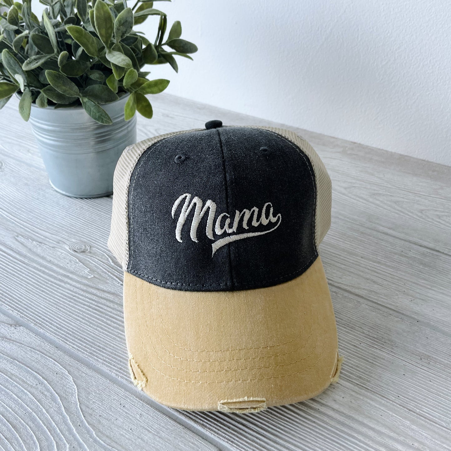 The Distressed Mama Hat in mustard and charcoal, featuring embroidery on a cotton front, is displayed on a light wood surface beside a small potted plant.