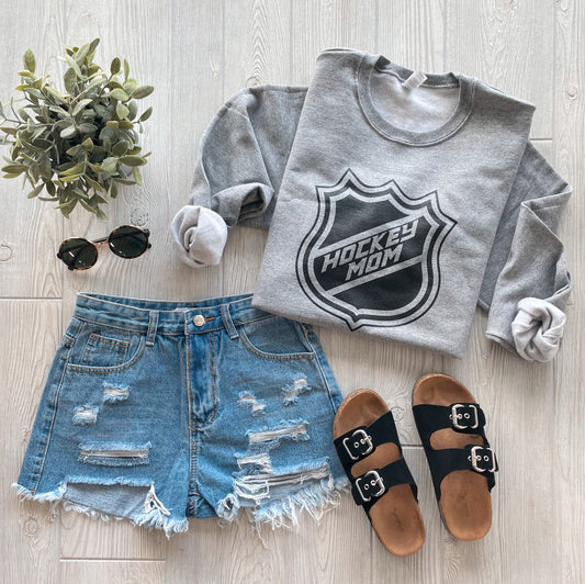 A flat lay from the Sports Mom Collection featuring the Hockey Mom • Gray Pullover, distressed denim shorts, black buckle sandals, round sunglasses, and a small green plant on a light wooden floor.