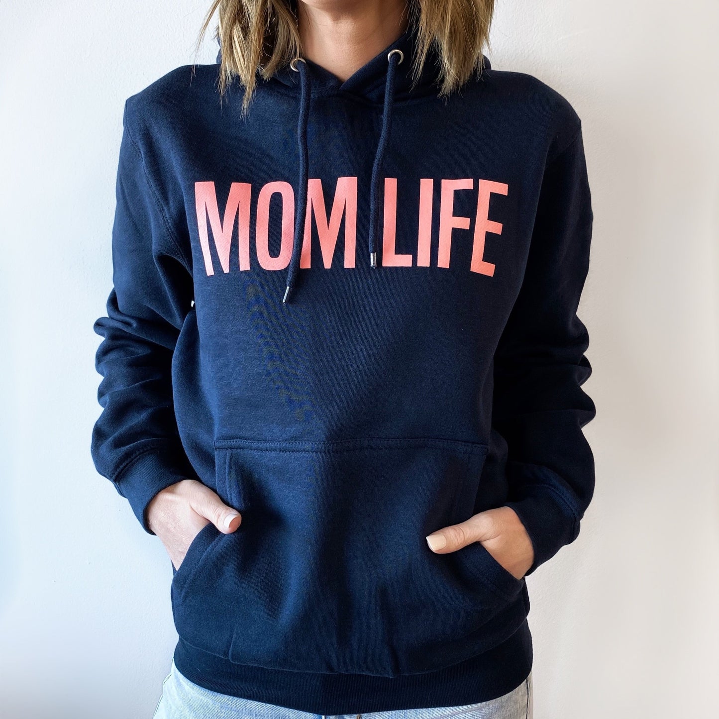 A person stands against a plain white background wearing the MOM LIFE • Navy Hoodie with large pink letters on the front and hands in the pocket.