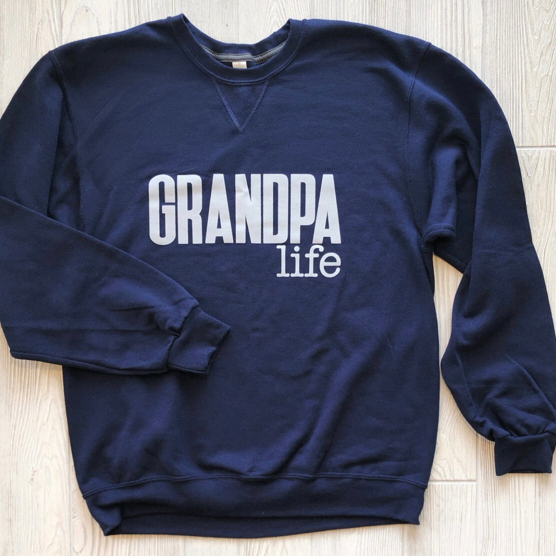 The Grandpa Pullover • Navy is a unisex navy blue sweatshirt with GRANDPA LIFE in bold white letters on the front, shown laid flat on a light wood floor.