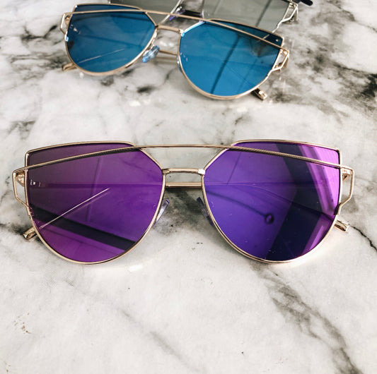Two pairs of Claire • Adult • More Colors! sunglasses with gold frames sit on marble—one pair with purple lenses in the foreground, another with blue lenses slightly out of focus in the background. Both offer UV protection.