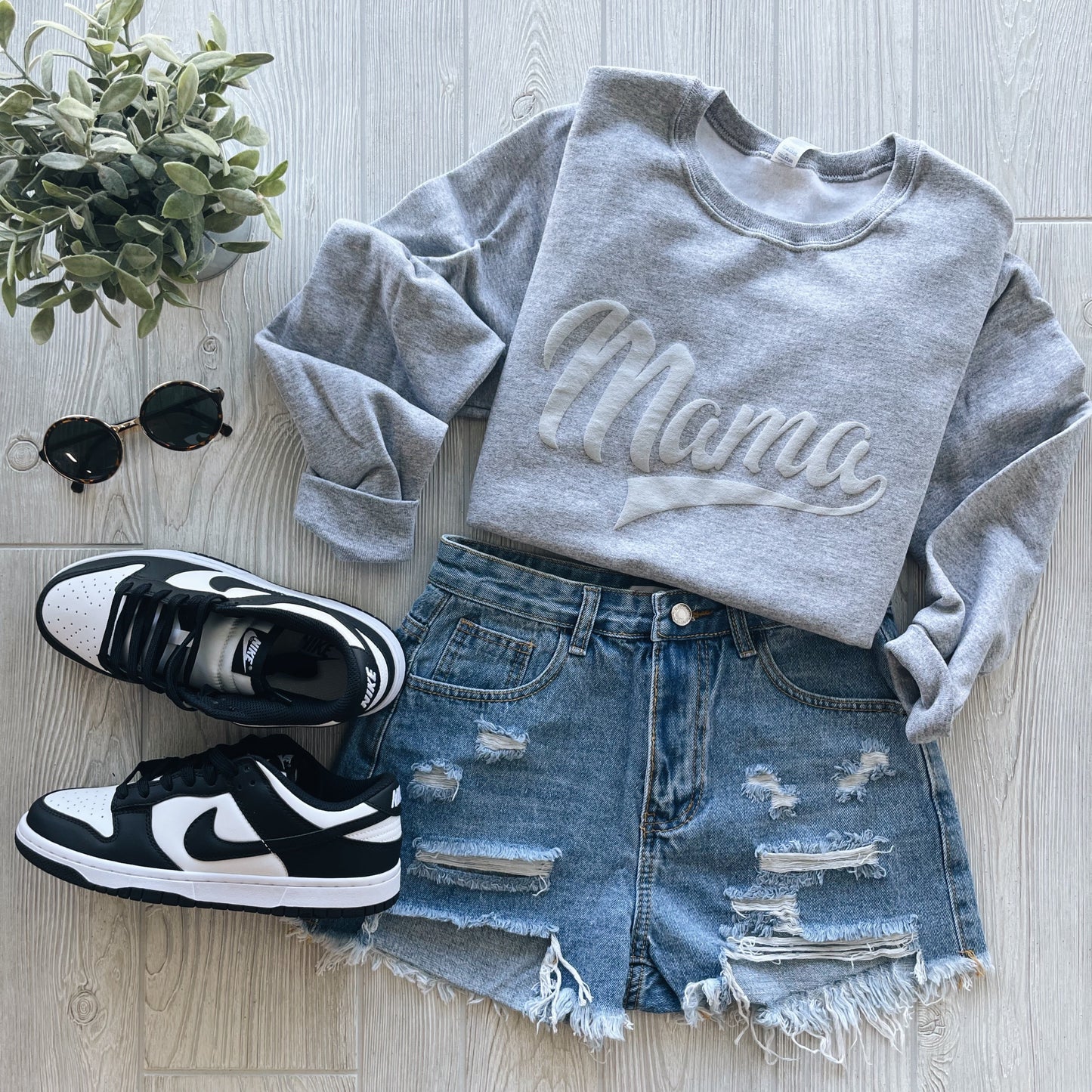 Flat lay of a casual outfit with the Mama Sporty • Gray Tonal Pullover, unisex fit, paired with distressed denim shorts, black and white Nike sneakers, sunglasses, and a potted plant on a light wood surface—ideal for cooler weather.