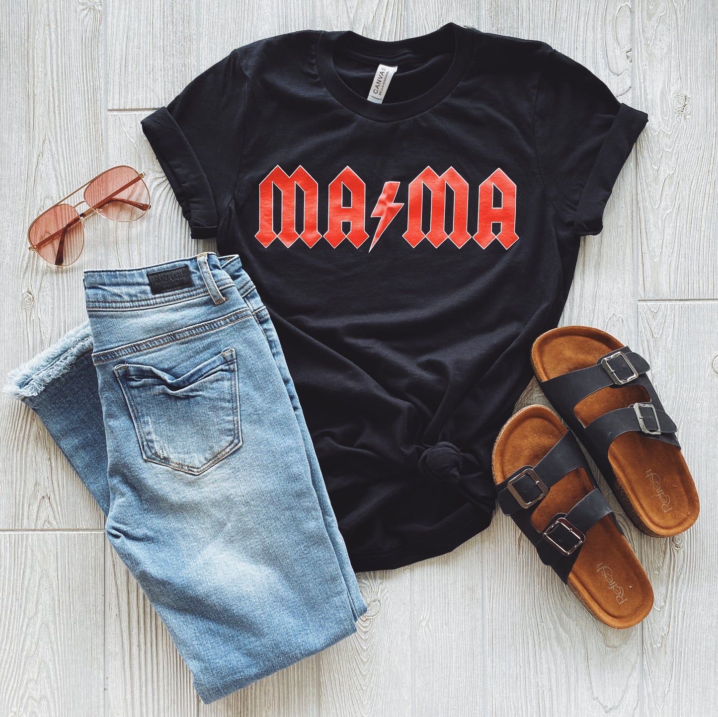 A flat lay features the MA⚡︎MA Rocker • Tee by Bella Canvas with an AC DC-inspired red and white design, paired with blue jeans, brown sandals with black straps, and rose gold sunglasses on a light wood floor.