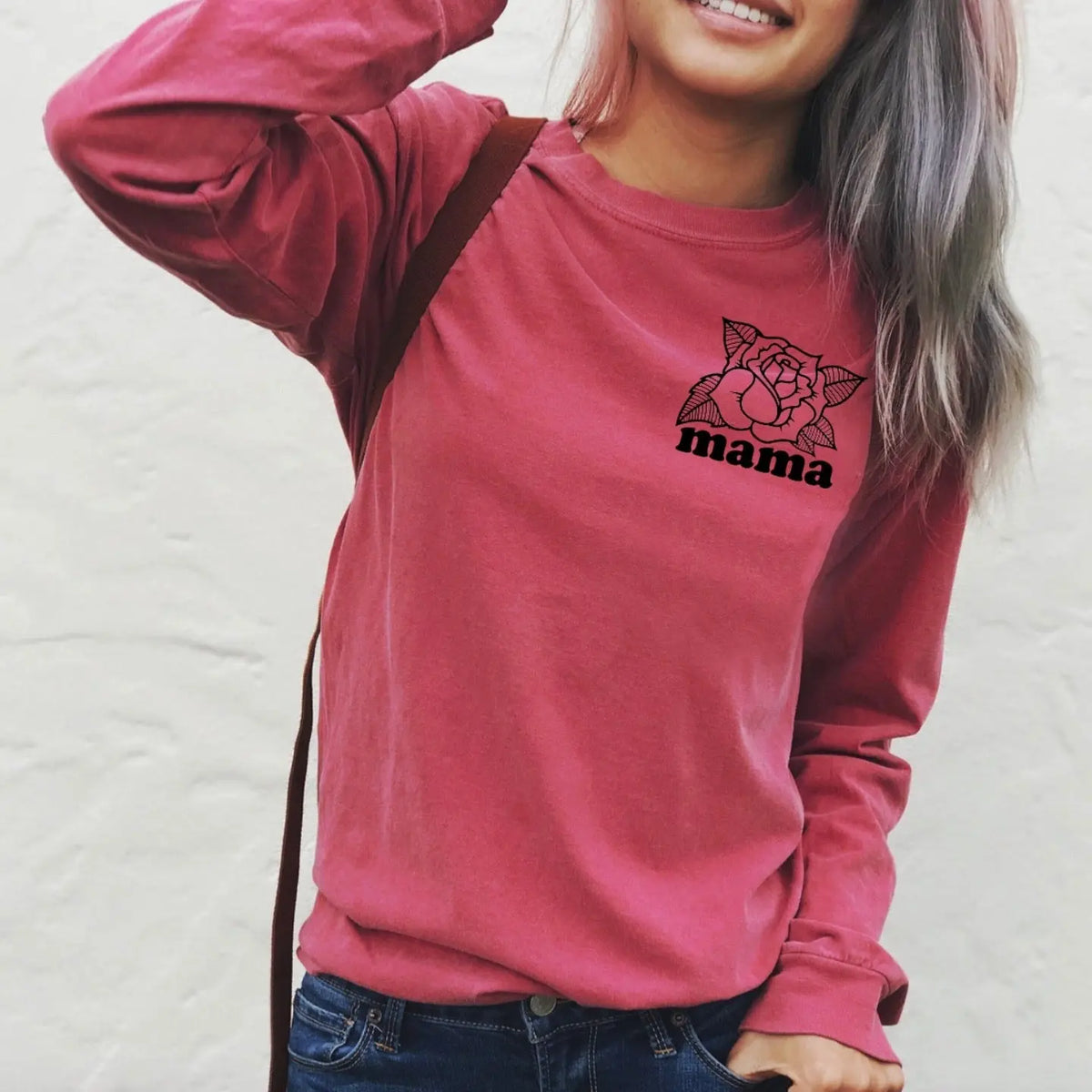 A woman smiles and touches her hair, wearing the Mama Rose • Long sleeve—a pink, garment-dyed shirt with a floral “mama” design—against a plain light background.