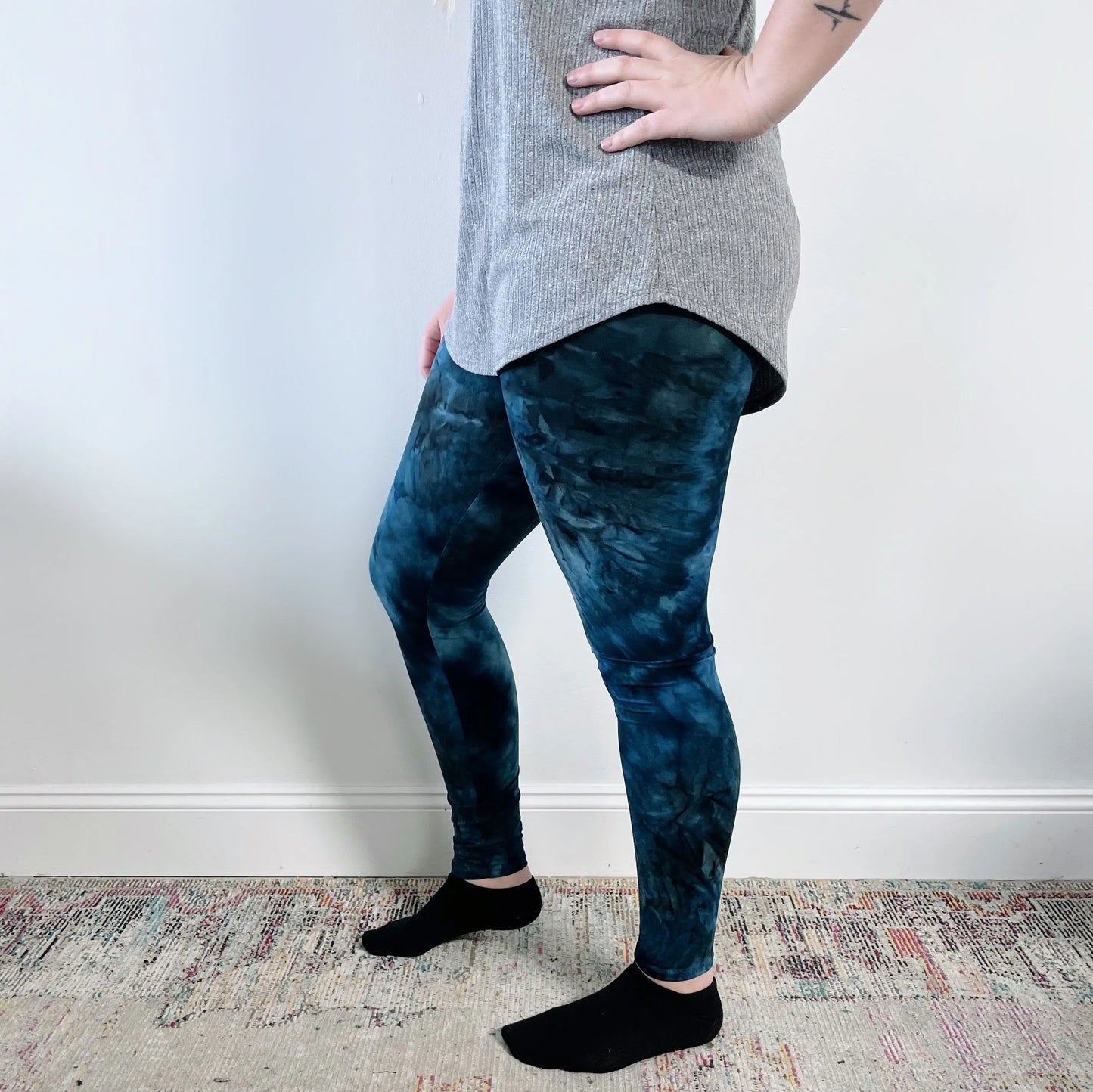 A person stands sideways with hand on hip, wearing a gray top and Extra Wide-Waist Leggings • Blue Dyed—super-soft leggings featuring a 5-inch waistband—paired with black socks, against a white wall and patterned rug.