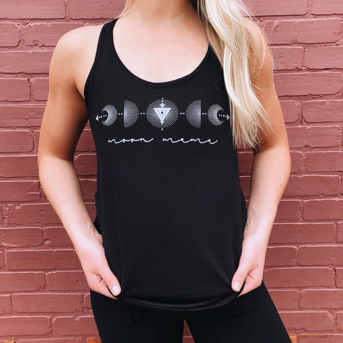A person with long blonde hair poses hands-in-pockets before a pink brick wall, wearing the Moon Mama • Slouchy Tank—a black women’s top with a flowy fit, moon phases, and “moon mama” text.