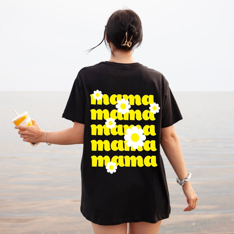 A woman stands by the water, holding a drink and wearing the Mama Daisy • Black Tee—a comfy unisex-fit Bella/Canvas shirt featuring “mama” in bold yellow with white daisies on the back.